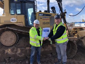 Two men shaking hands exchanging a certificate in front of a bull dozer